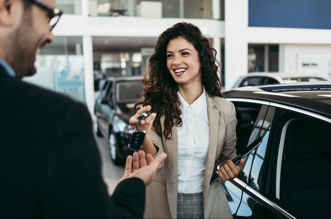 A imagem mostra uma mulher recebendo as chaves do carro que acabou de comprar.