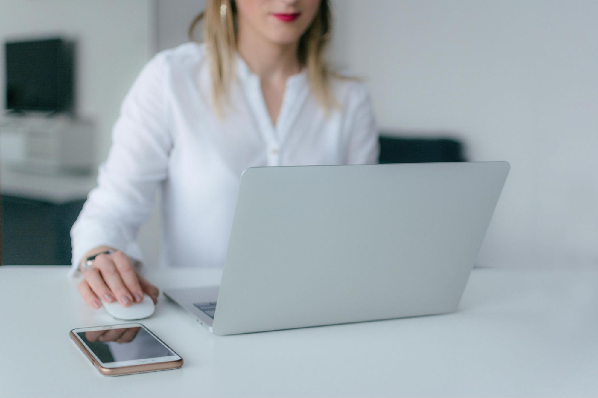 Mulher usando camisa social branca e usando seu laptop na cor cinza. Em sua mesa, um smartphone.