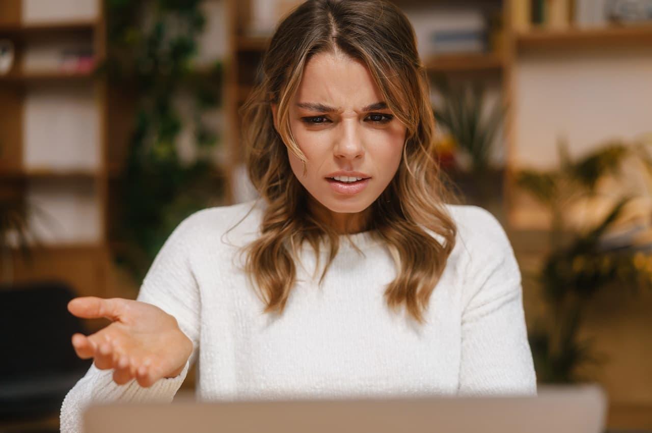 Mulher com expressão confusa ou preocupada, olhando para um laptop. Ela está em um ambiente com prateleiras ao fundo, vestindo um suéter branco.