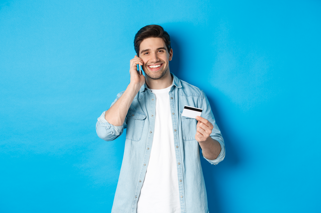 Homem sorridente com uma camiseta branca e camisa jeans segurando um cartão de crédito e falando ao telefone, em frente a um fundo azul.