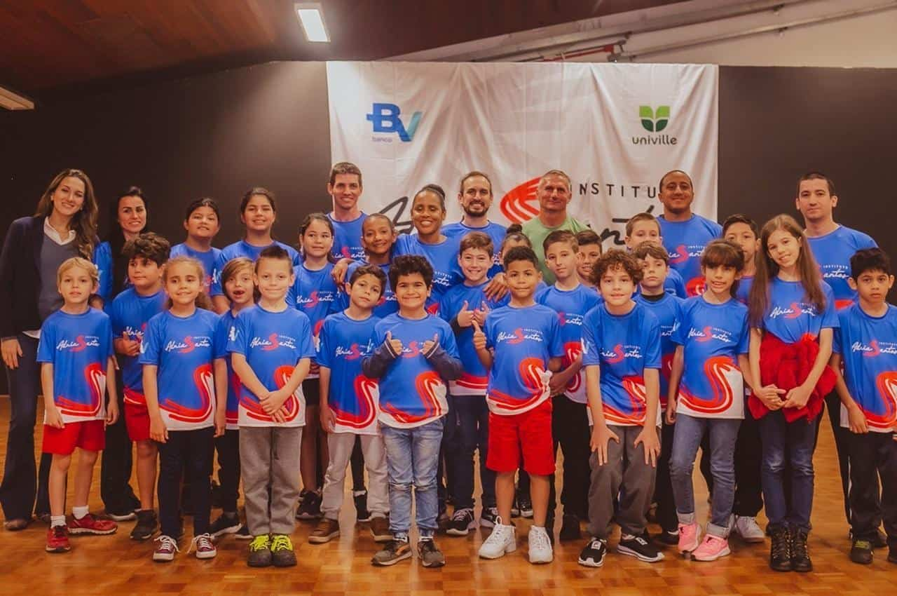 Grupo de crianças sorridentes posando para uma foto em grupo ao lado dos professores de práticas esportivas. Todos vestem camiseta azul da marca banco BV.