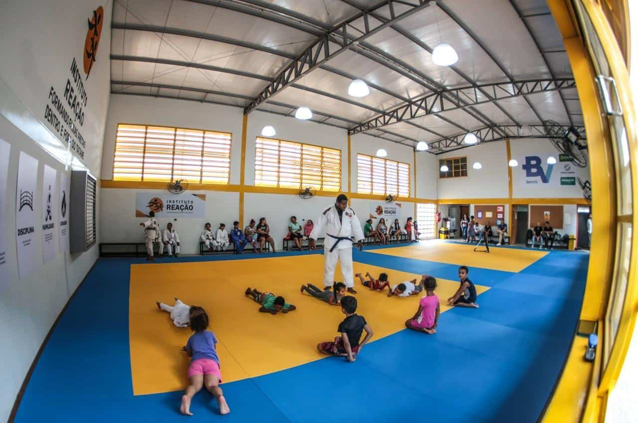 Professor de judô em pé no tatame azul e ensinando crianças sobre a prática esportiva de judô. As pessoas estão em um ginásio escolar.