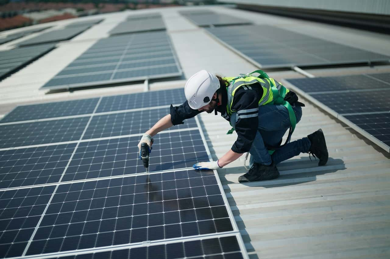 Engenheiro de energia solar agachado fazendo a instalação de energia solar. Ele veste colete, luvas e capacete de segurança e usa uma furadeira para instalar placa solar