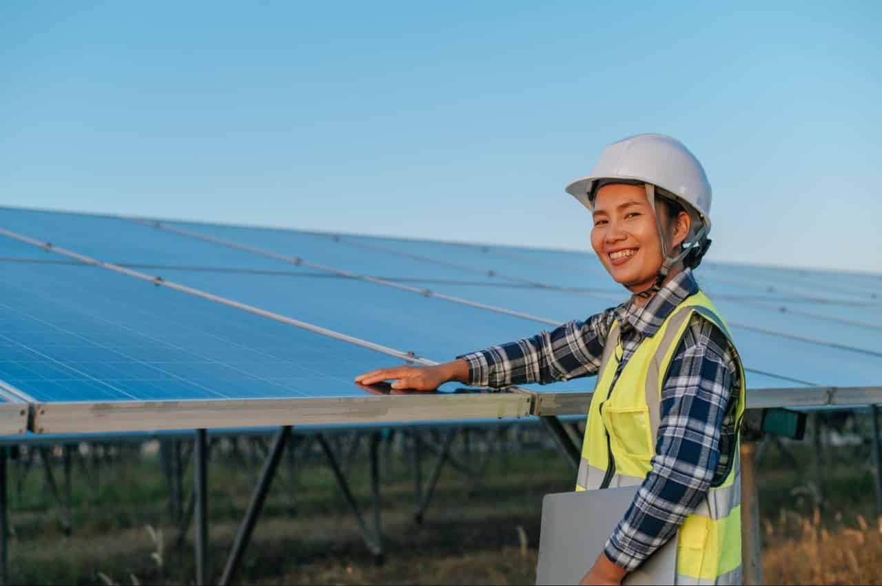 Mulher sorridente com prancheta, capacete branco e roupa de proteção amarela encostando a palma da mão na placa solar.