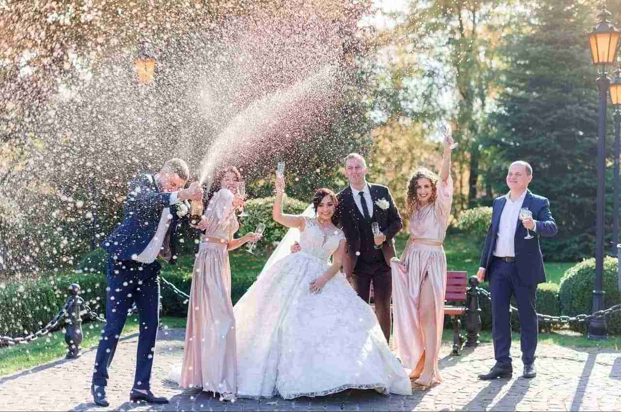 Casal sorridente de recém-casados (mulher com vestido de noiva e homem com um terno preto) comemorando com seus convidados de casamento e jogando champanhe ao redor.