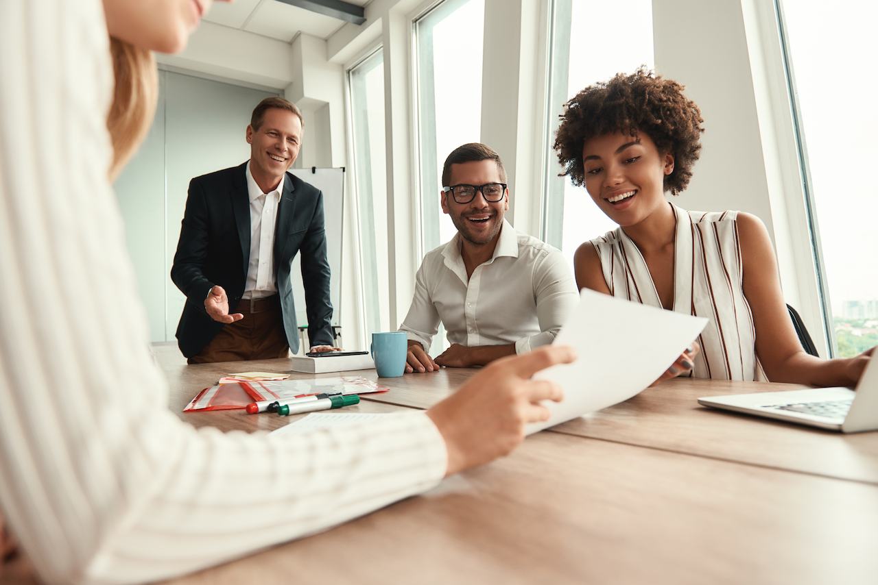 A imagem mostra quatro pessoas em uma mesa de reunião. Uma delas está mostrando um papel e as outras estão o analisando e sorrindo.