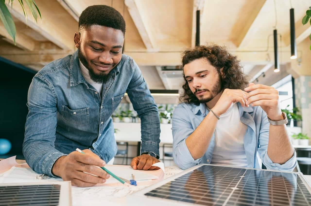 Dois homens conversando sobre qual é o melhor inversor de energia solar. Eles observam uma placa solar e fazem anotações no caderno.