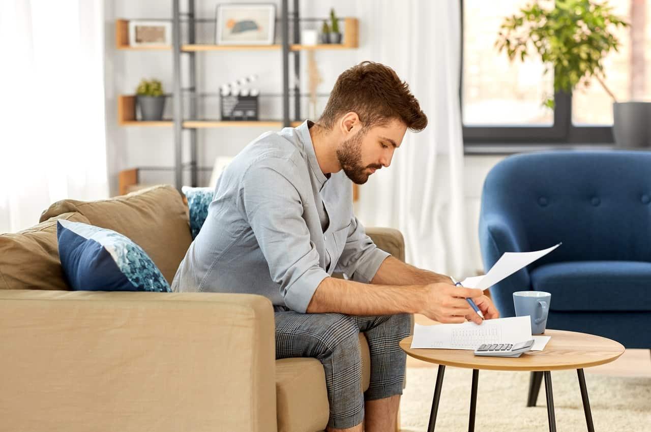 Homem sentado no sofá segura folhas de papel e uma calculadora com a expressão concentrada. Ele veste calça xadrez cinza e camiseta jeans.