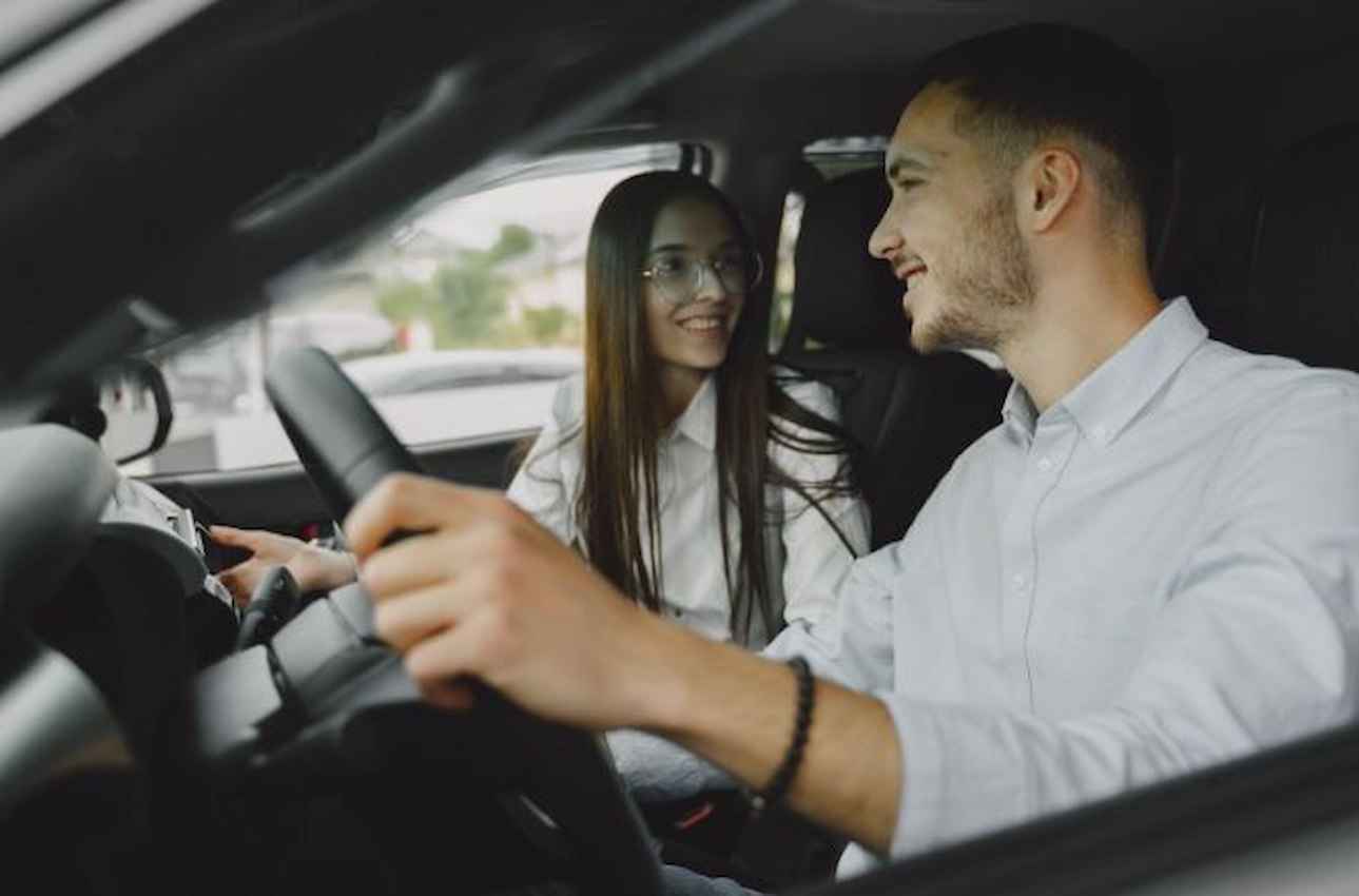 A imagem mostra um casal, dentro de seu carro. Eles estão sorridentes.