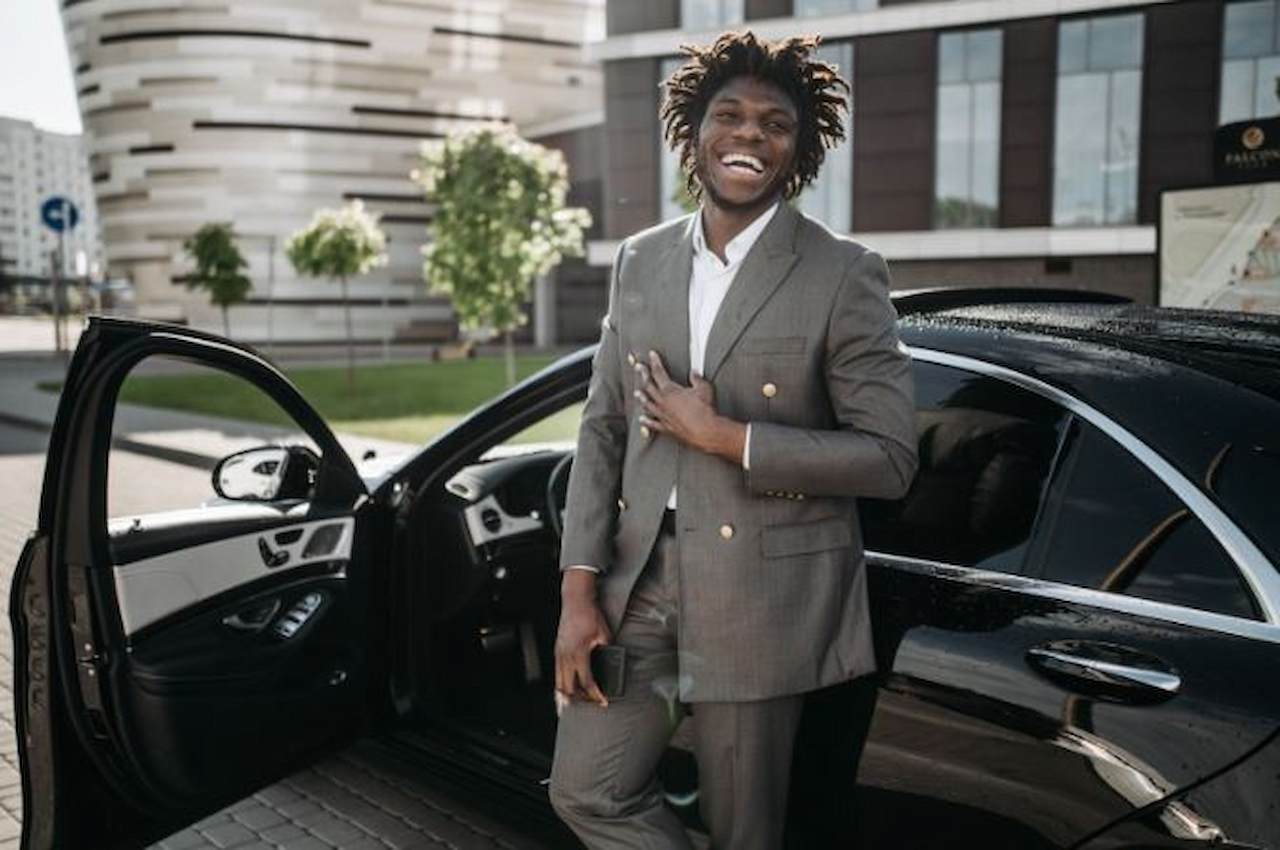 A imagem mostra um homem a frente de seu carro, que está com a porta do motorista aberta. Ele está segurando um celular e está sorridente.