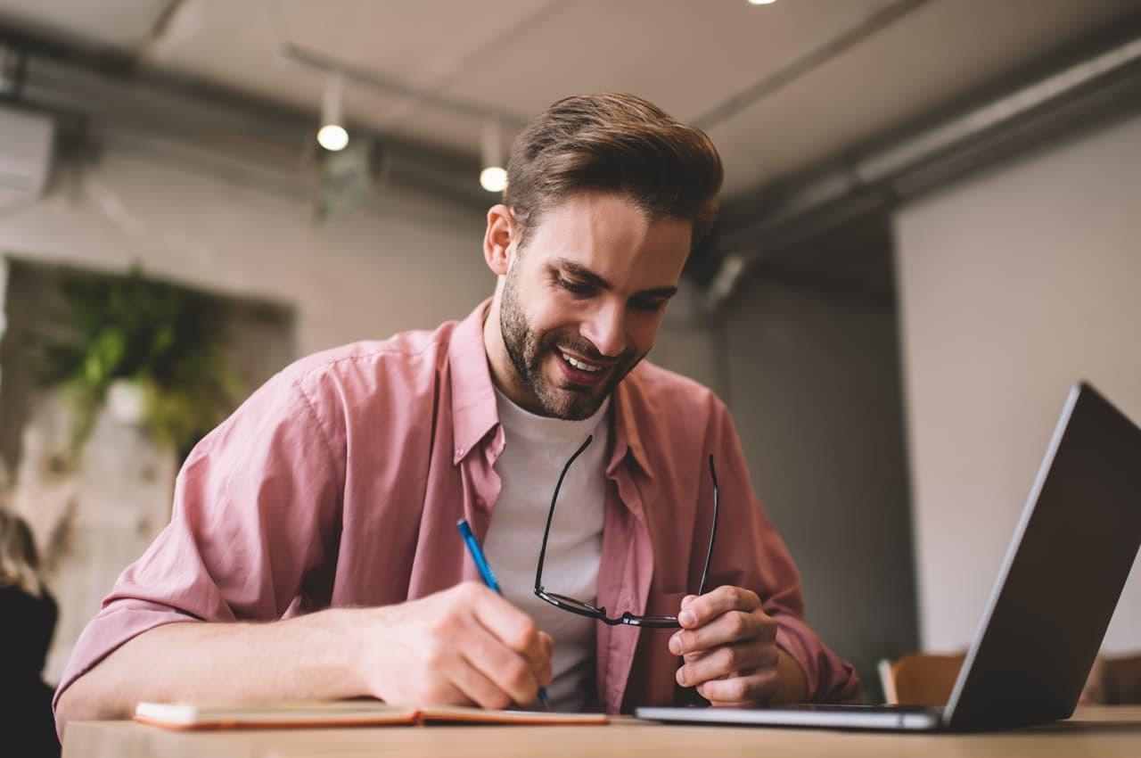 Homem sorrindo enquanto escreve e segura seus óculos. Ele está em frente a um notebook, fazendo anotações sobre o LTV de empréstimo.