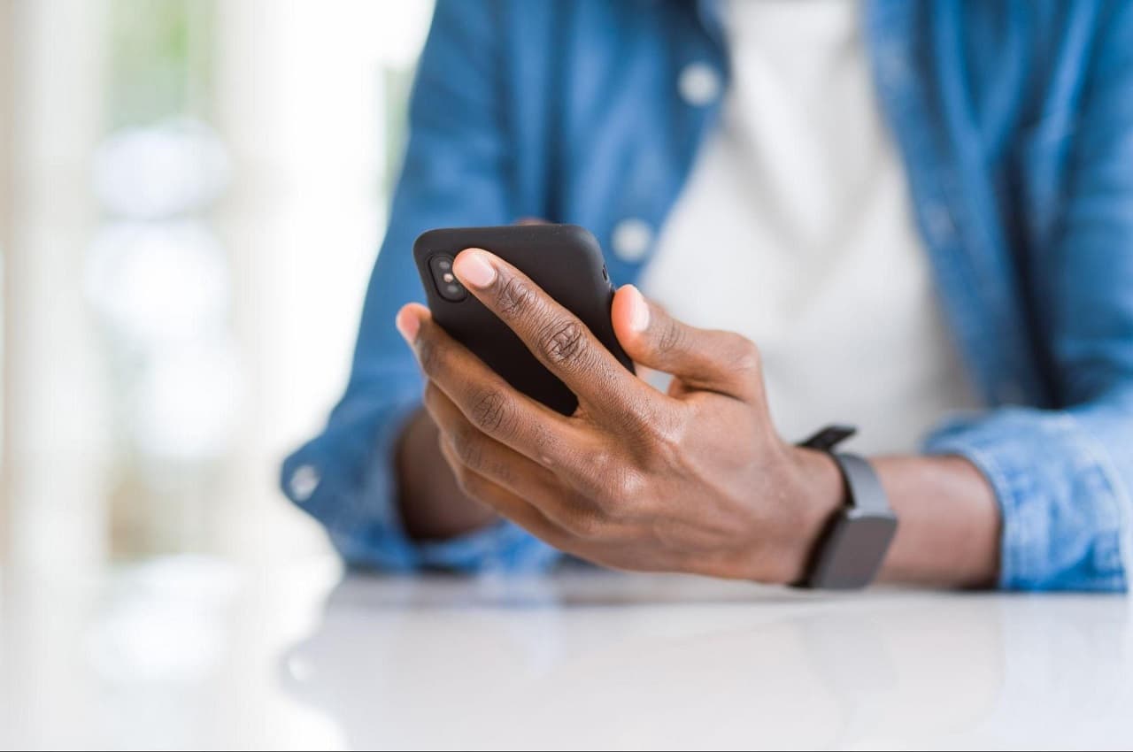 Homem (somente seus braços e mãos aparecem na imagem) digitando no celular. Ele veste camisa branca básica, jaqueta jeans e está sentado na mesa de sua casa.