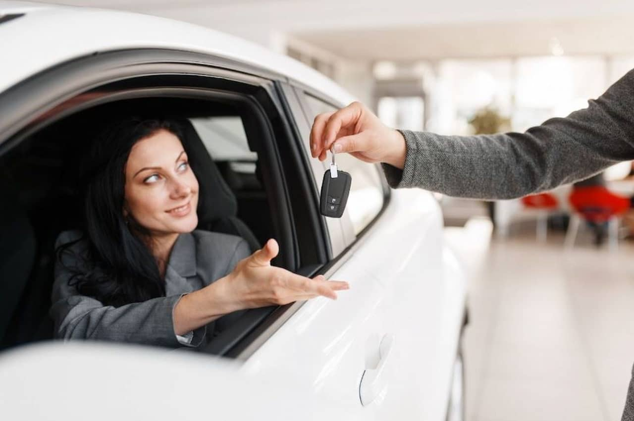 Mulher com expressão sorridente recebendo as chaves do seu carro novo pelo vendedor.