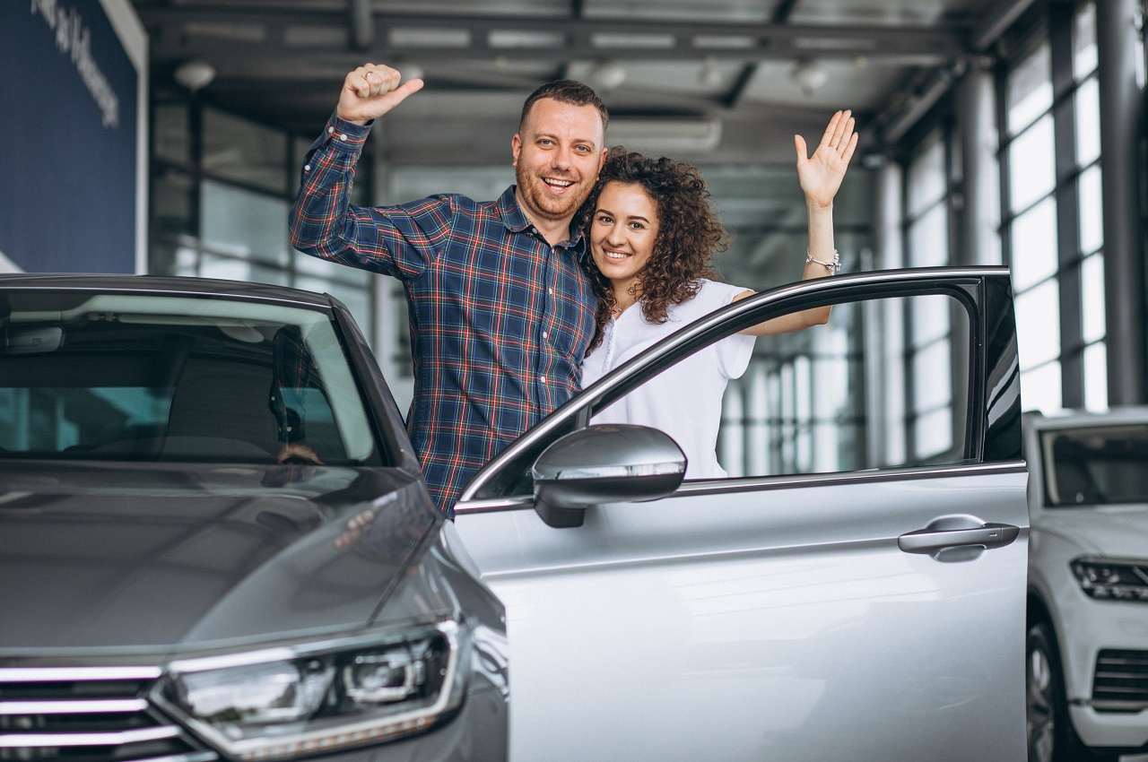 Homem e mulher abraçados fazendo joianha com as mãos e em frente ao seu primeiro carro.