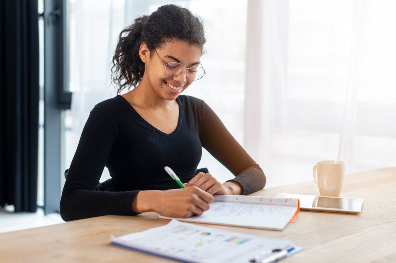 A imagem mostra uma mulher sentada à uma mesa, fazendo anotações em um caderno.