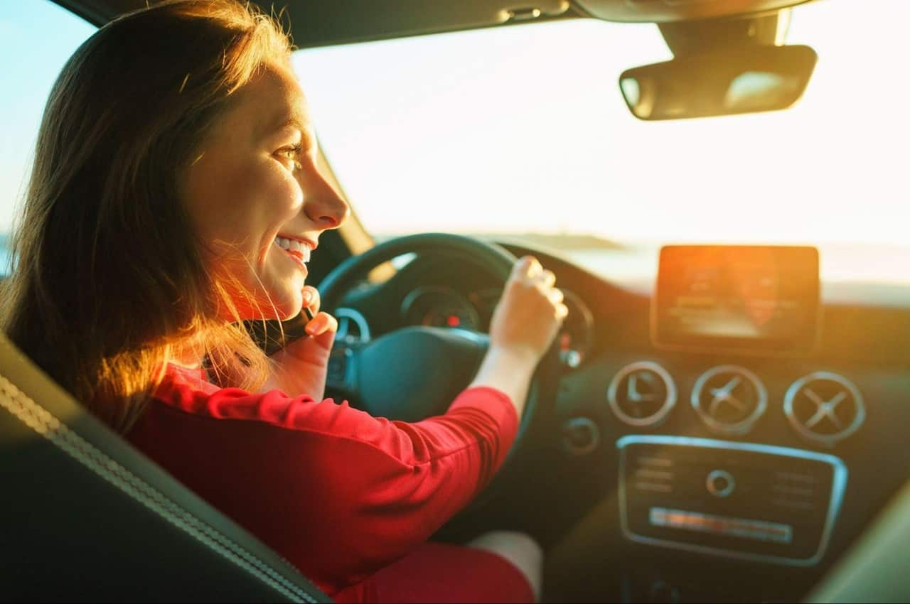 Mulher sorridente de cabelos castanhos dirigindo um carro e olhando para o lado. O sol bate em seu rosto, iluminando o ambiente.