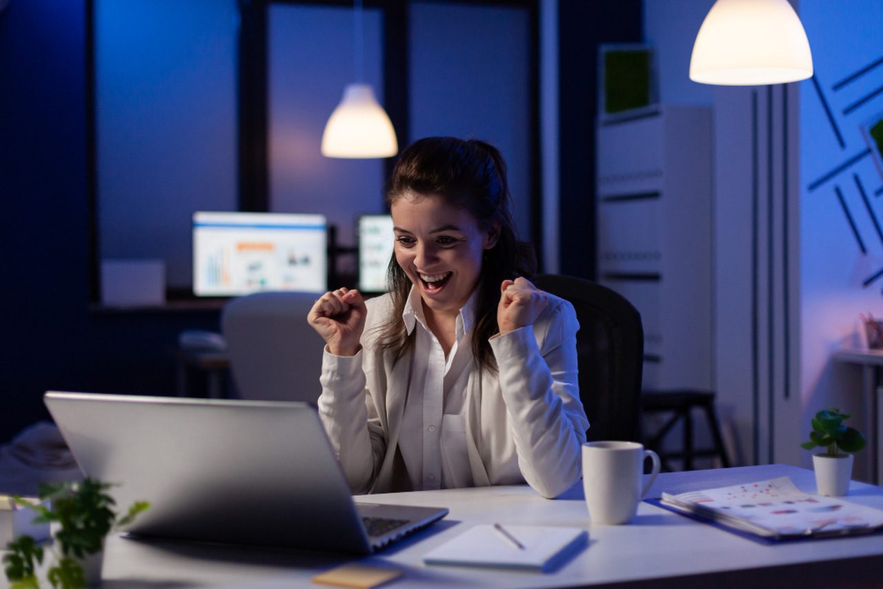 Mulher sorridente vestindo uma camisa branca e lendo ótimas notícias online no computador.