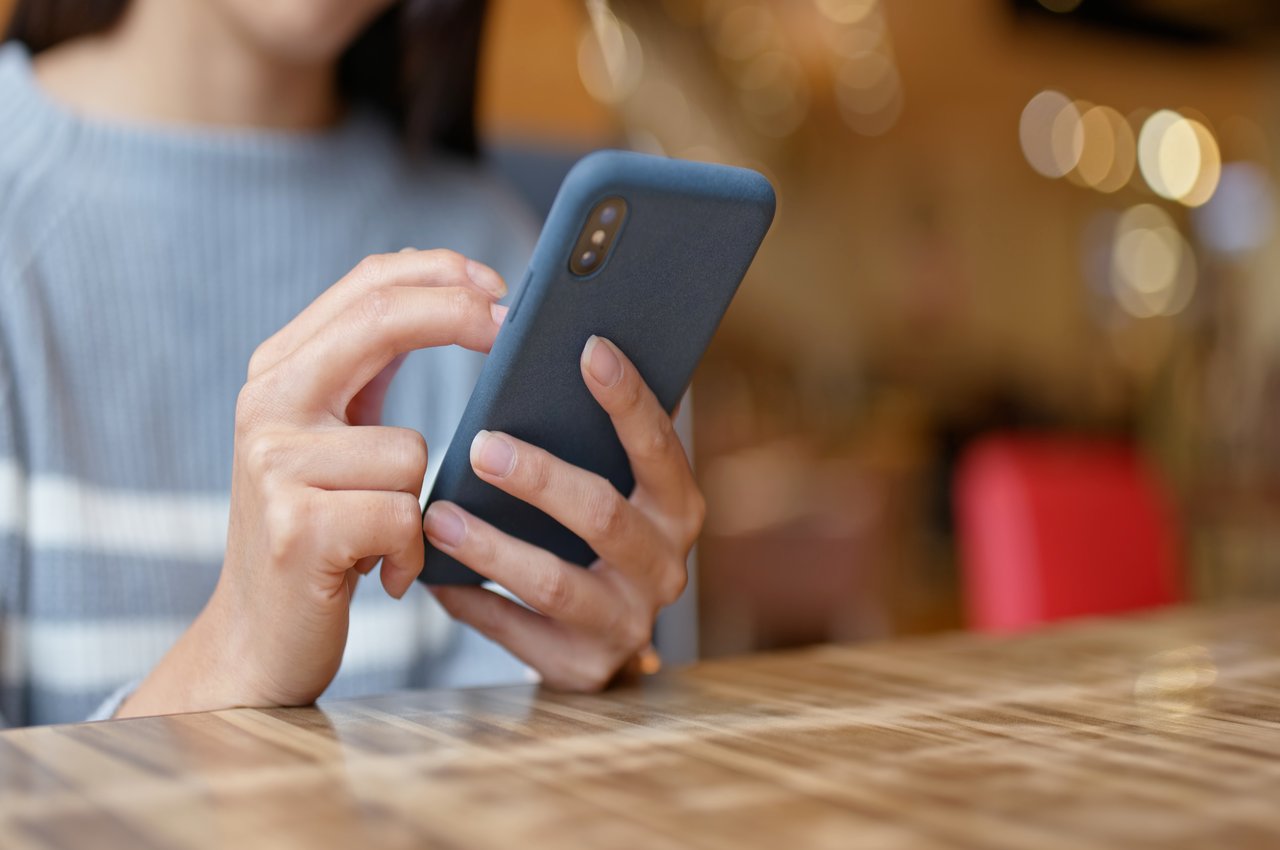 Foco em celular azul sendo utilizado por uma pessoa (seu rosto não aparece na imagem). A pessoa está sentada na mesa de um restaurante.