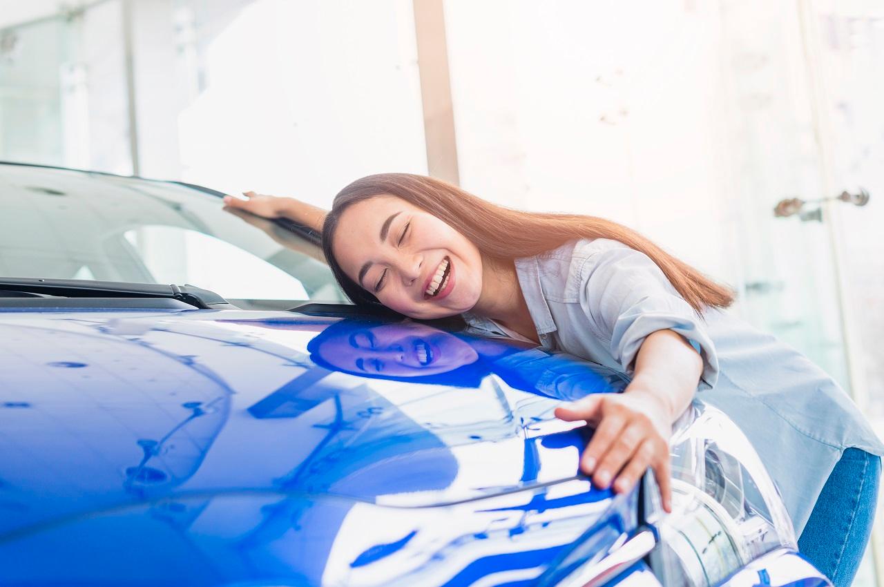 Mulher com expressão sorridente e de olhos fechados, abraçando o capo do seu novo carro híbrido na concessionária. 