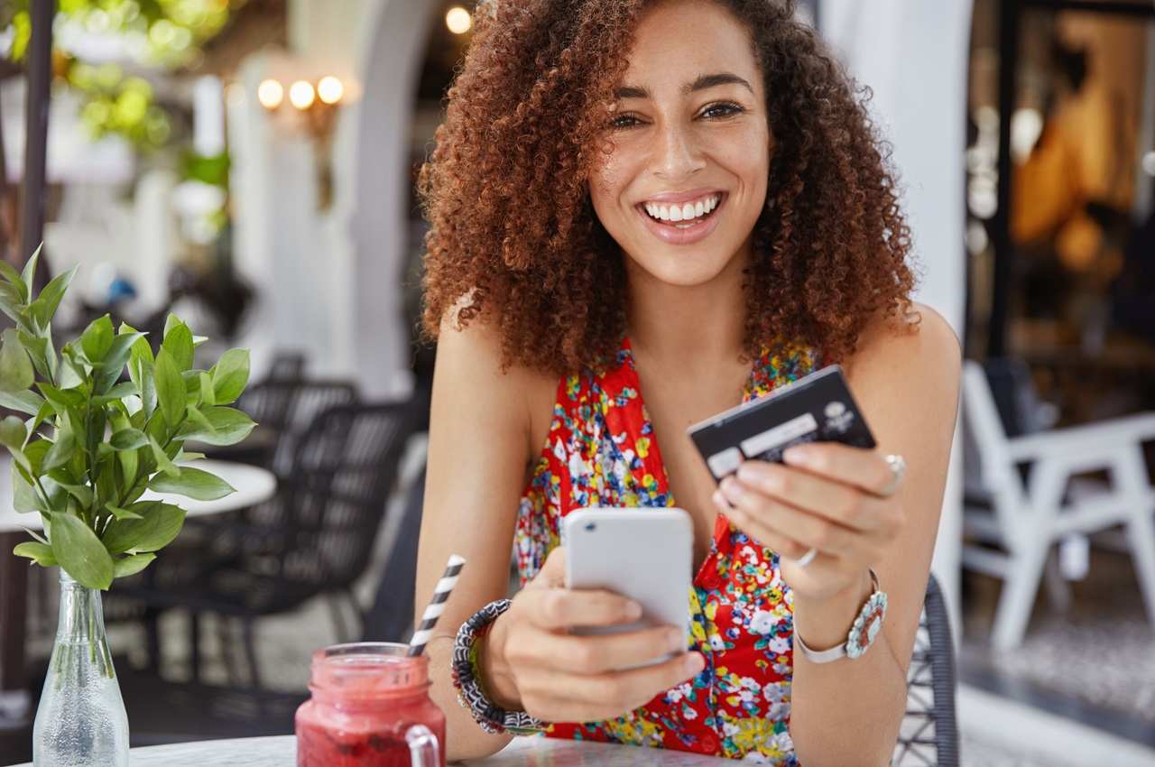 A imagem mostra uma mulher sorridente em um restaurante, com um suco em sua mesa. Ela está com o celular em uma mão e o cartão de crédito em outra.