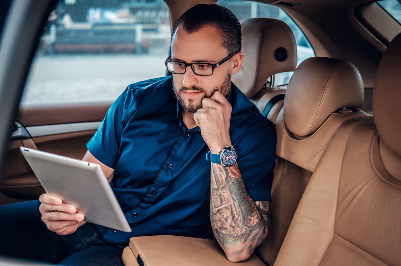 Homem com óculos sentado no banco traseiro de um carro. Ele está segurando um tablet e com um expressão concentrada.