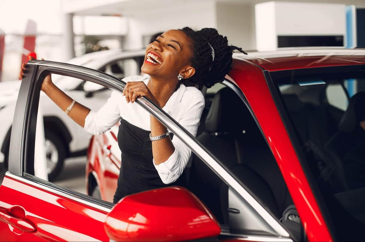 A imagem mostra uma mulher se apoiando na porta de um carro vermelho. Ela está sorridente.