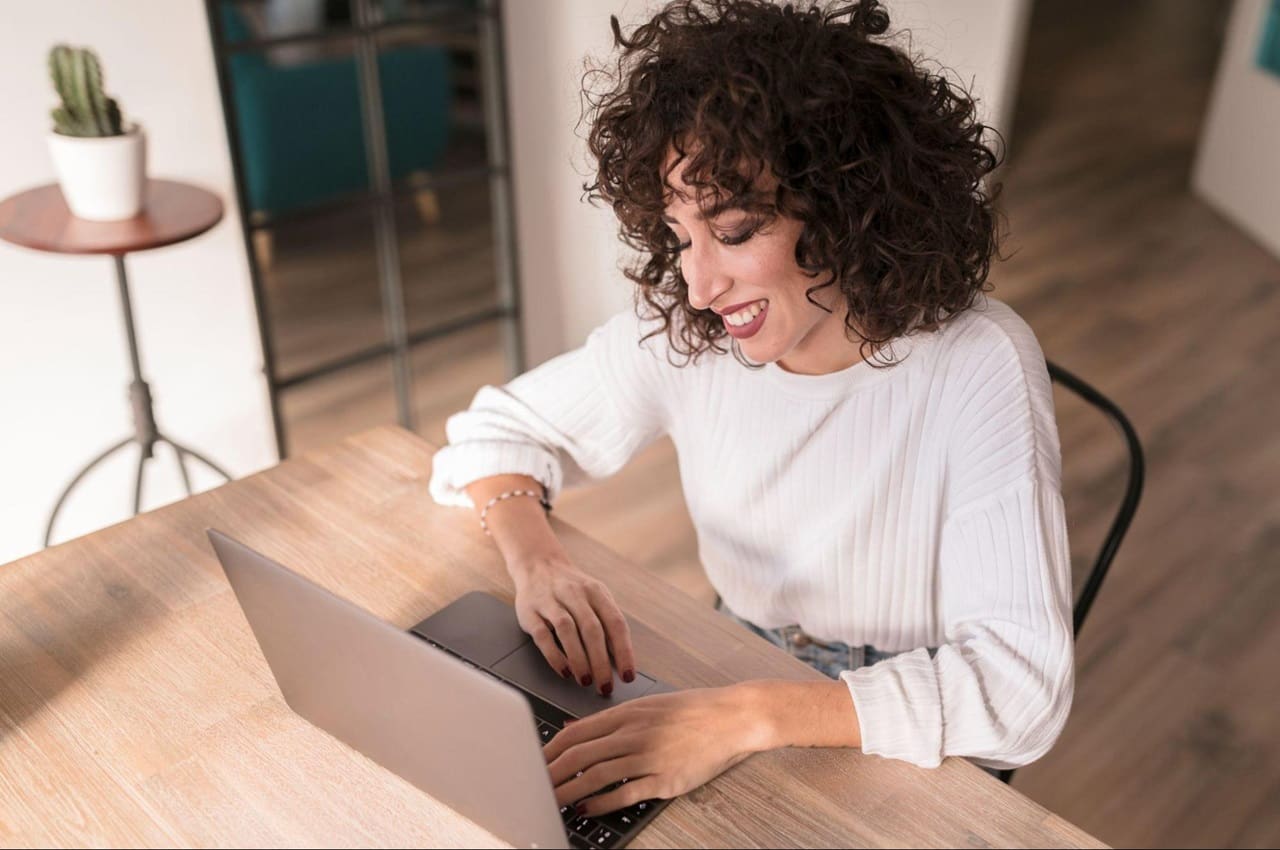 Mulher sorridente digita no laptop e veste camisa de mangas longas brancas.