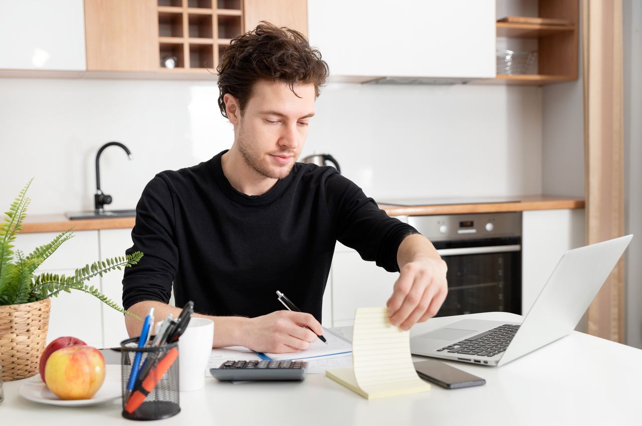 A imagem mostra um homem sentado em uma mesa, em uma cozinha. Ele está usando seu laptop e algumas anotações em um papel.