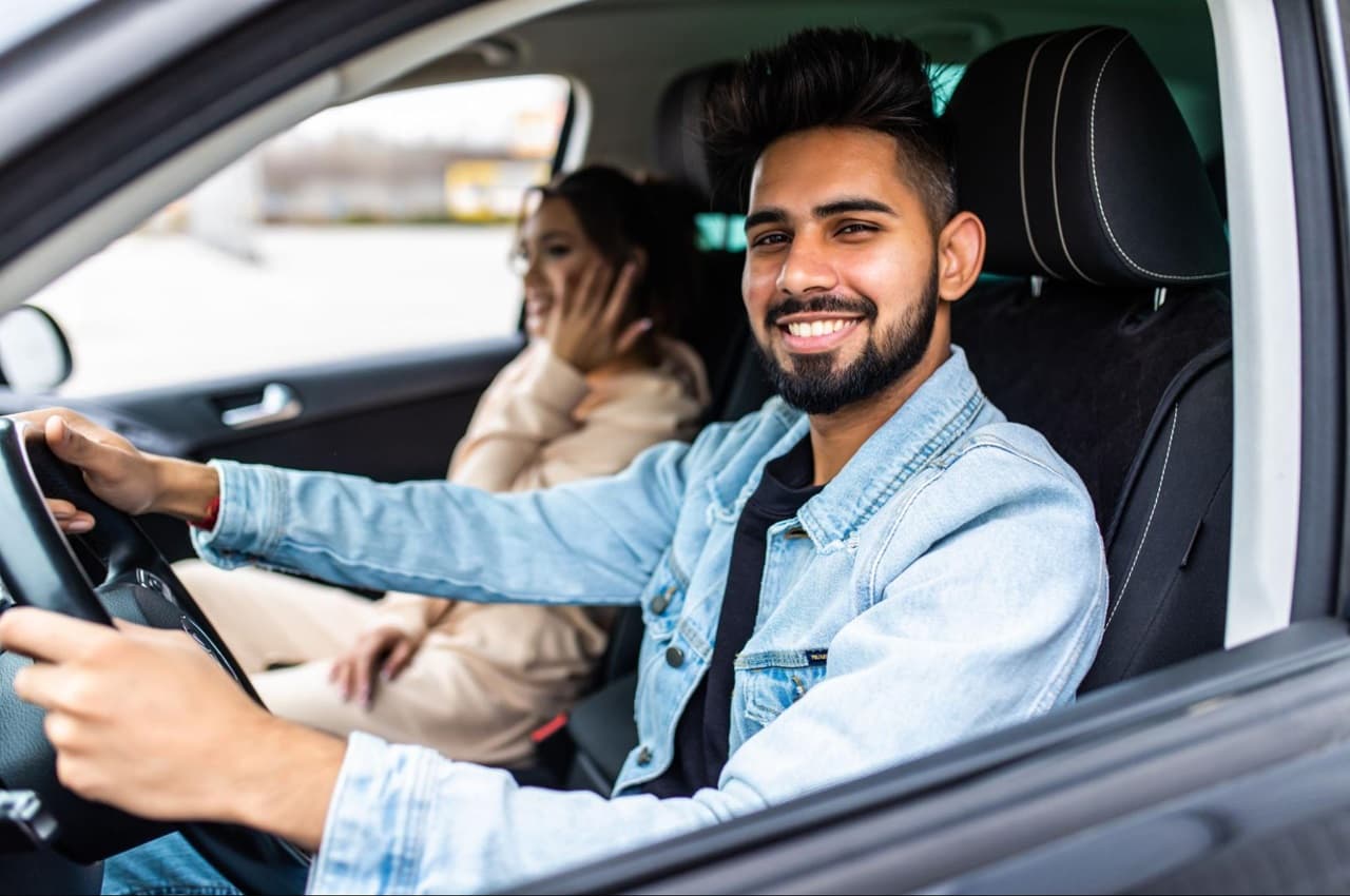 Homem sorridente e de jaqueta jeans dirigindo um veículo. No banco de passageiro está uma mulher também sorridente.