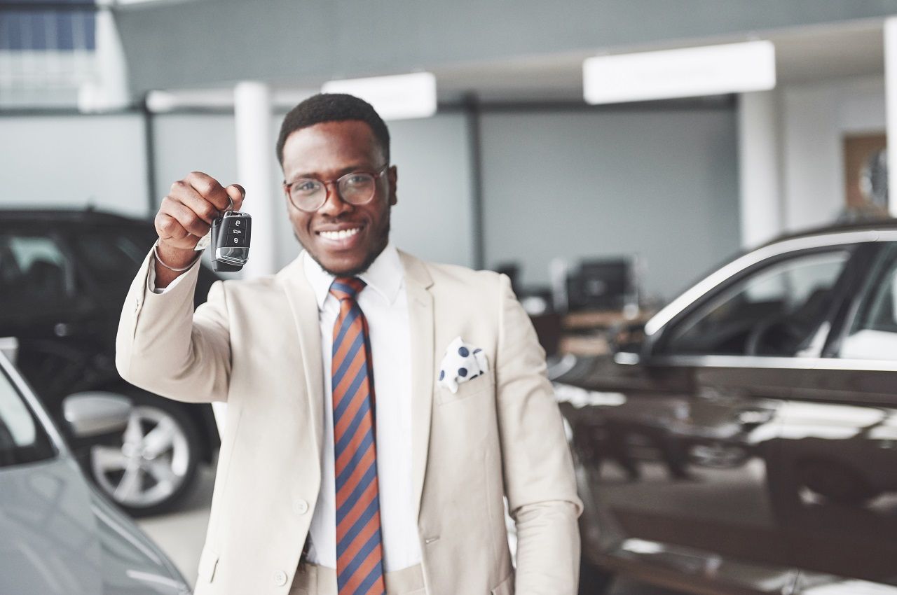 Homem sorridente, vestindo conjunto de terno e segurando uma chave de carro na concessionária.