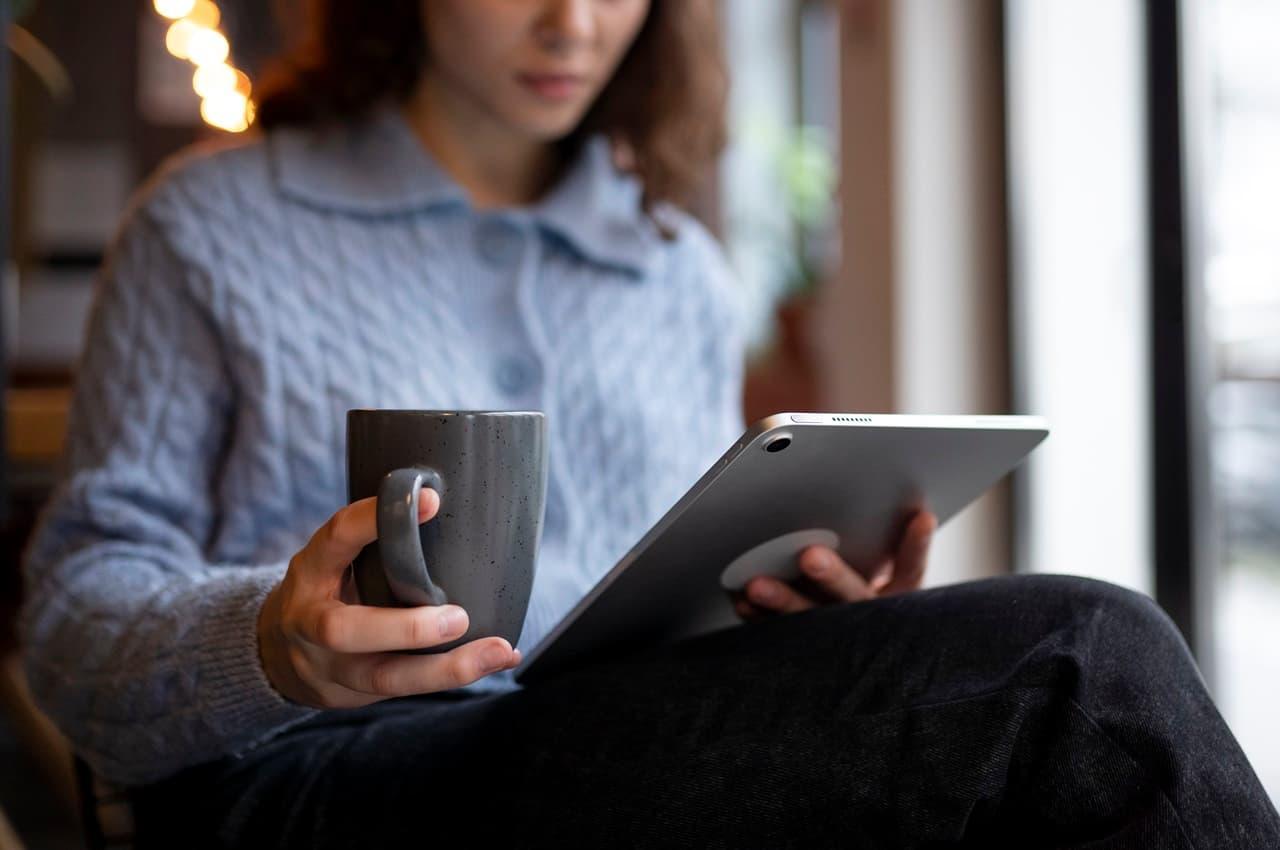 Mulher veste suéter azul de tricô, segura xícara de café e usa tablet para consultar renavam do veículo.