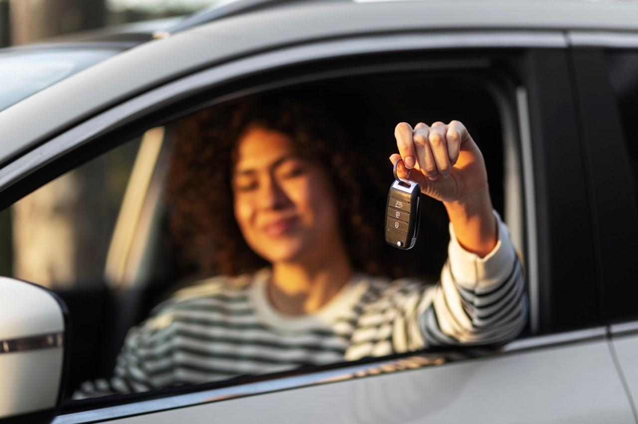 Mulher com semblante feliz no interior de um carro. Ela está sentada no banco do motorista, com o vidro aberto, e segurando as chaves.