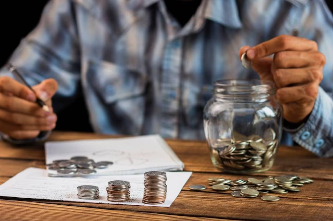 A imagem mostra um homem usando uma camisa xadrez, contando, anotando e colocando moedas em um cofre.