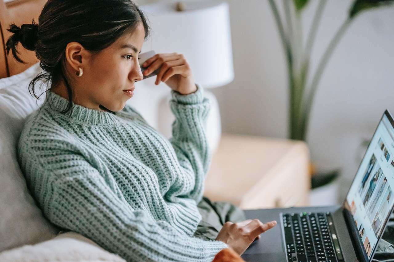 A imagem mostra uma mulher sentada em um sofá, usando seu laptop para fazer compras.