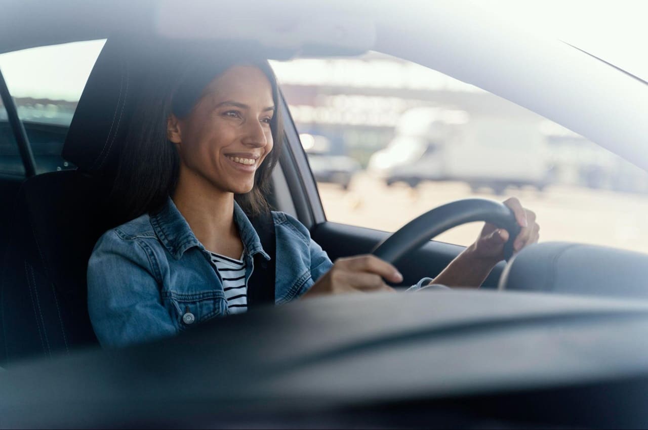 Mulher sorridente de cabelos castanhos lisos, jaqueta jeans e blusa listrada, usa cinto de segurança e dirige veículo em avenida movimentada.