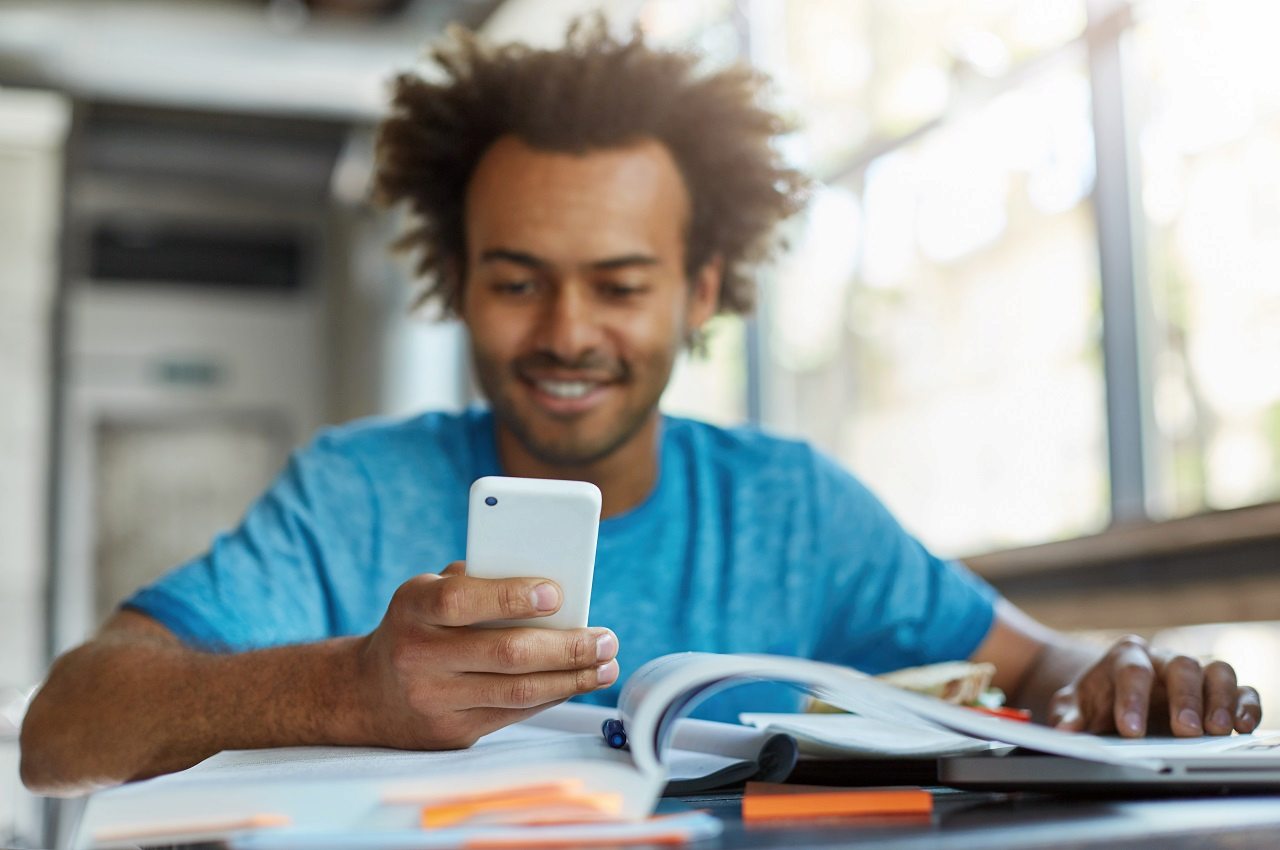 Homem sorridente, vestindo blusa básica azul e digitando no celular.