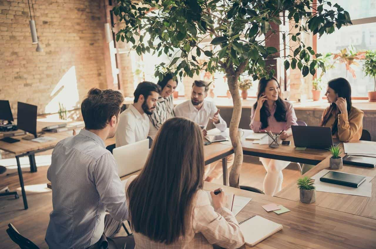 Grupo de pessoas sentadas e sorridentes em um escritório e ao redor de uma árvore, indicando que seguem os critérios ESG.