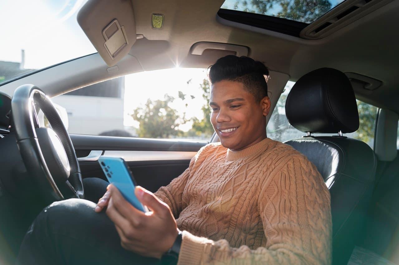 Motorista de aplicativo sorridente segura celular azul para escolher próxima corrida e está sentado no banco de motorista do veículo. 