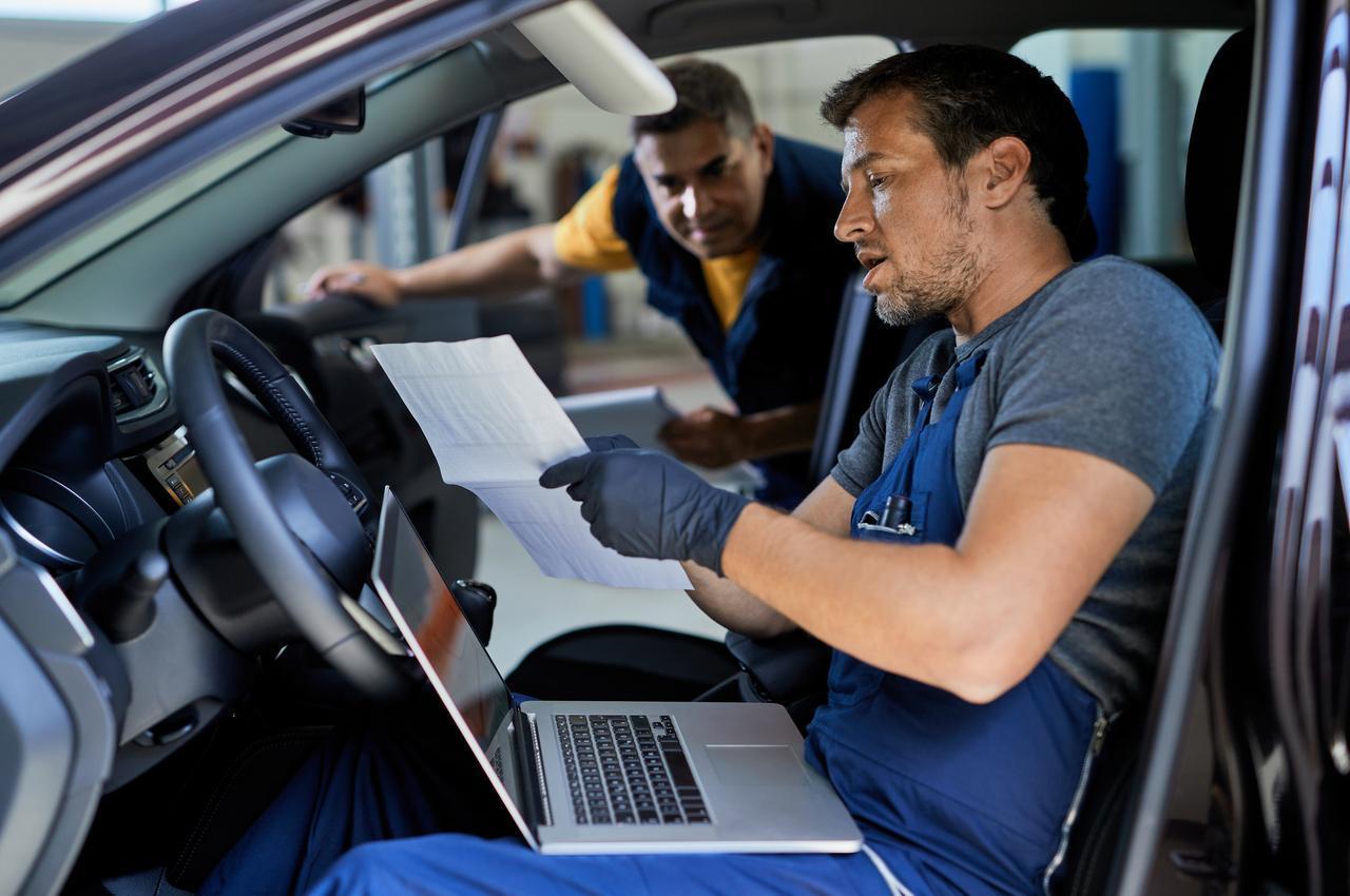 Dois mecânicos realizando vistoria veicular dentro de um carro, um deles segurando um documento e usando um notebook.