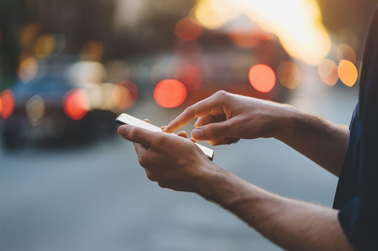 Pessoa (seu rosto não aparece na imagem) clicando com dedo indicador na tela de um celular. Ao fundo, tem luzes borradas da rua 