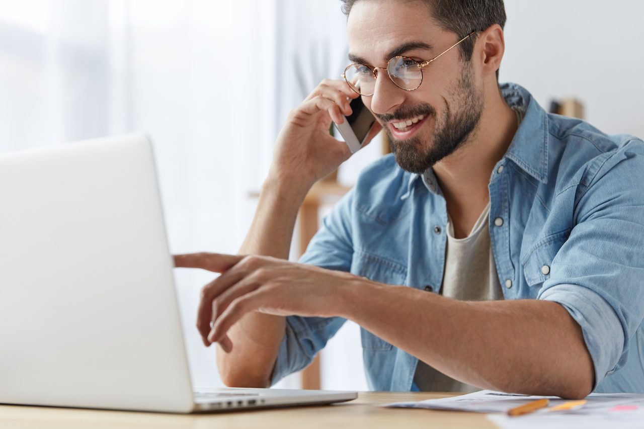 A imagem mostra um homem falando ao celular e apontando para a tela do seu laptop.