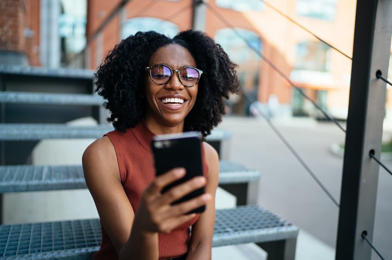 A imagem mostra uma mulher sorridente, ao ar livre, usando seu telefone.