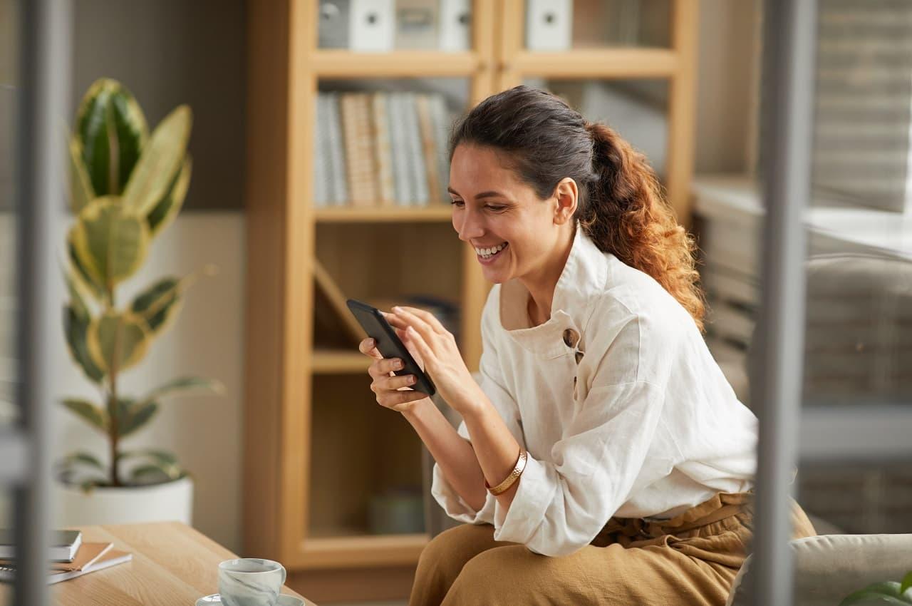 A imagem mostra uma mulher sentada, sorridente e usando seu celular.