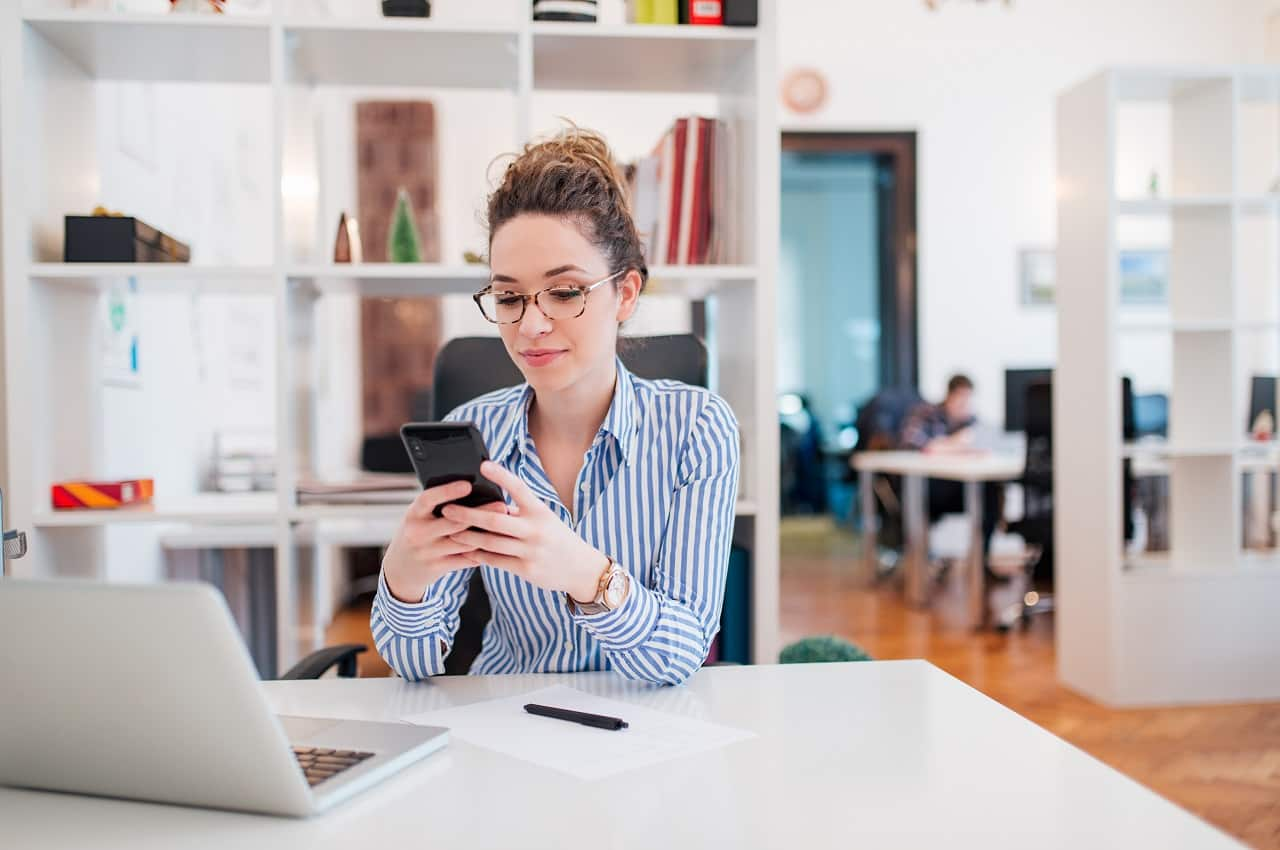 Mulher com expressão concentrada, cabelos cacheados presos e blusa social listrada. Ela digita no celular e está sentada na mesa do escritório.