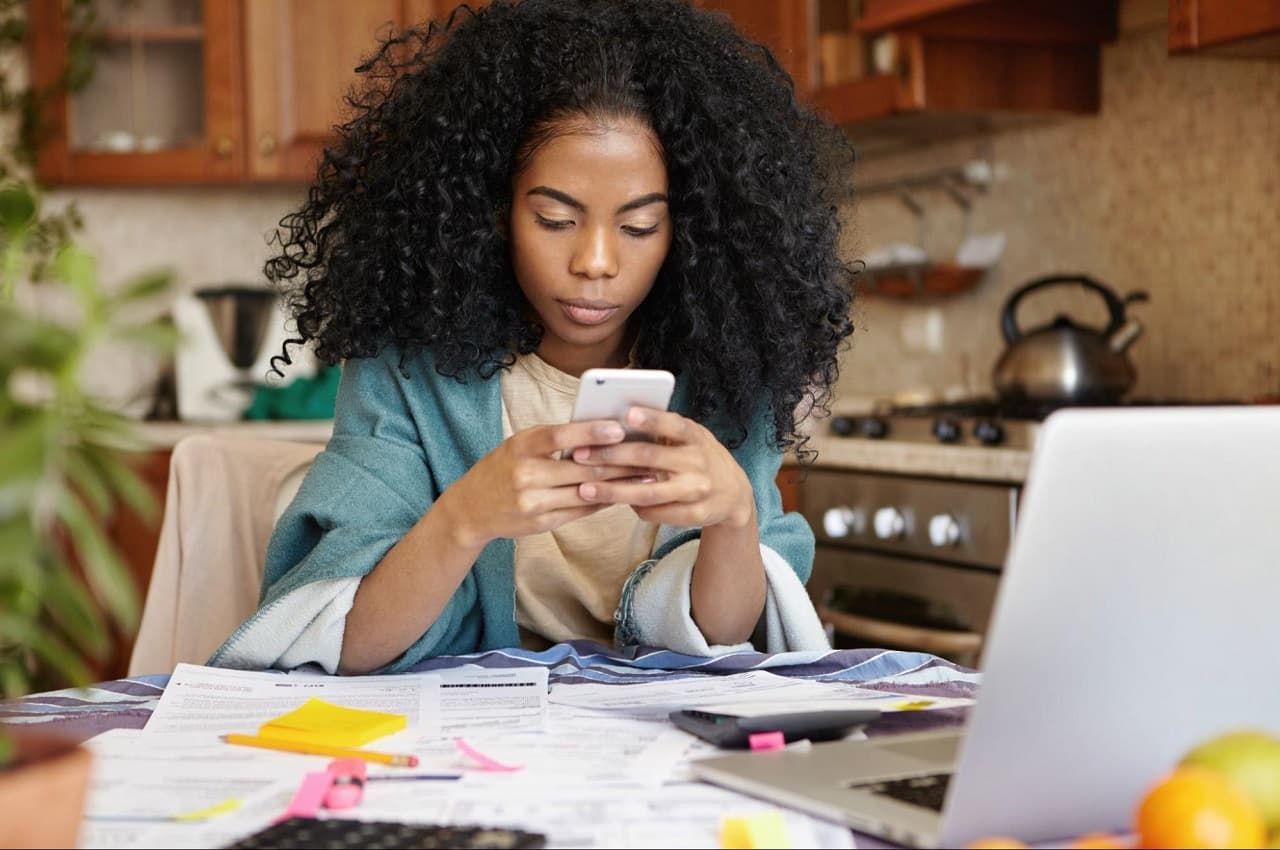 A imagem mostra uma mulher, sentada a uma mesa com vários papéis e um laptop. Ela está usando seu celular.