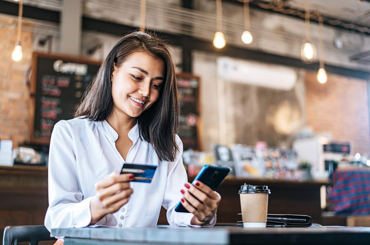 A imagem mostra uma mulher em um estabelecimento, tomando um café. Ela está se preparando para fazer o pagamento com seu cartão por aproximação.