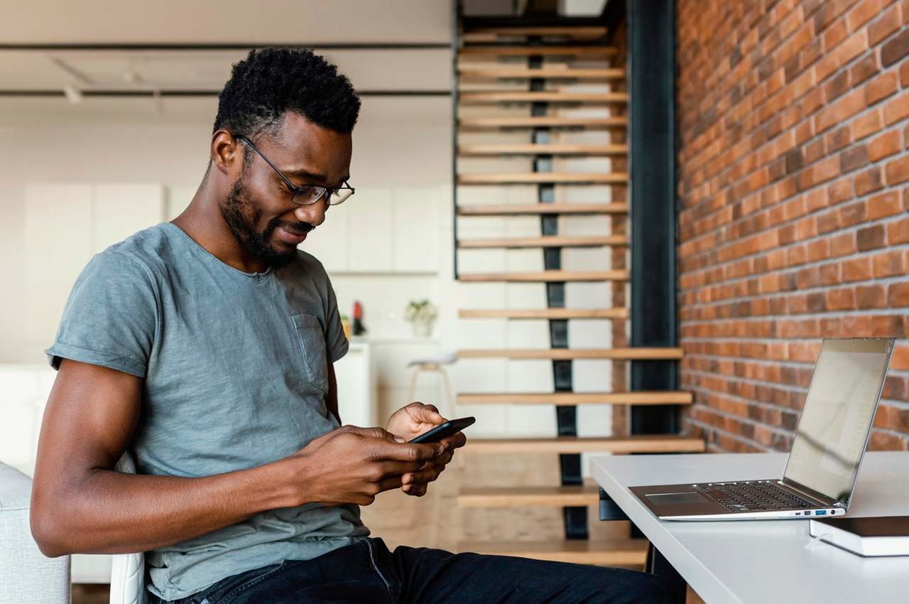 A imagem mostra um homem sorridente usando seu celular e seu laptop.