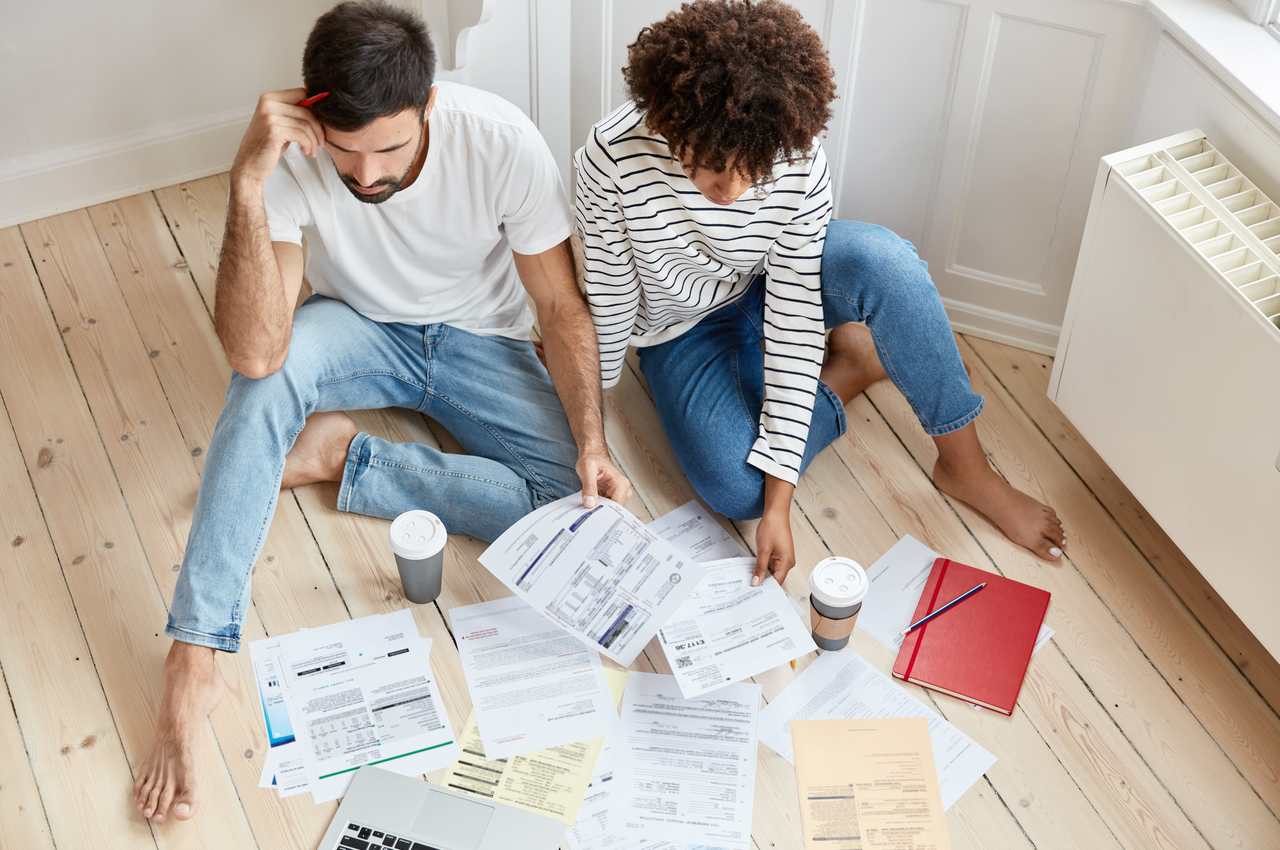 Homem e mulher com expressões concentradas sentados um do lado do outro no chão de uma casa, olhando para contas de início do ano espalhadas pelo chão.