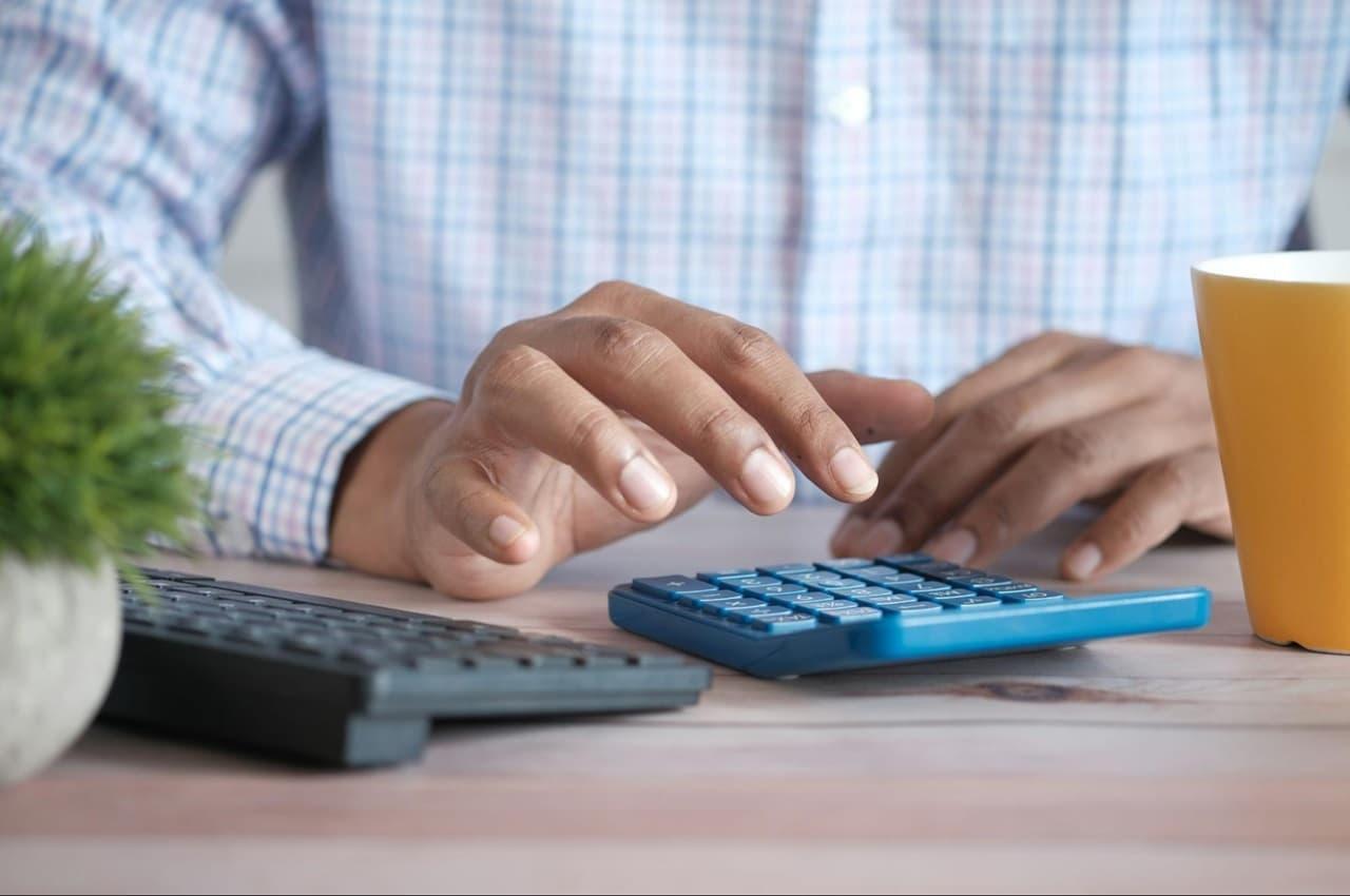 Foco em mãos calculando IPVA de carro comprado direto com proprietário. A pessoa usa blusa xadrez azul e uma calculadora simples.