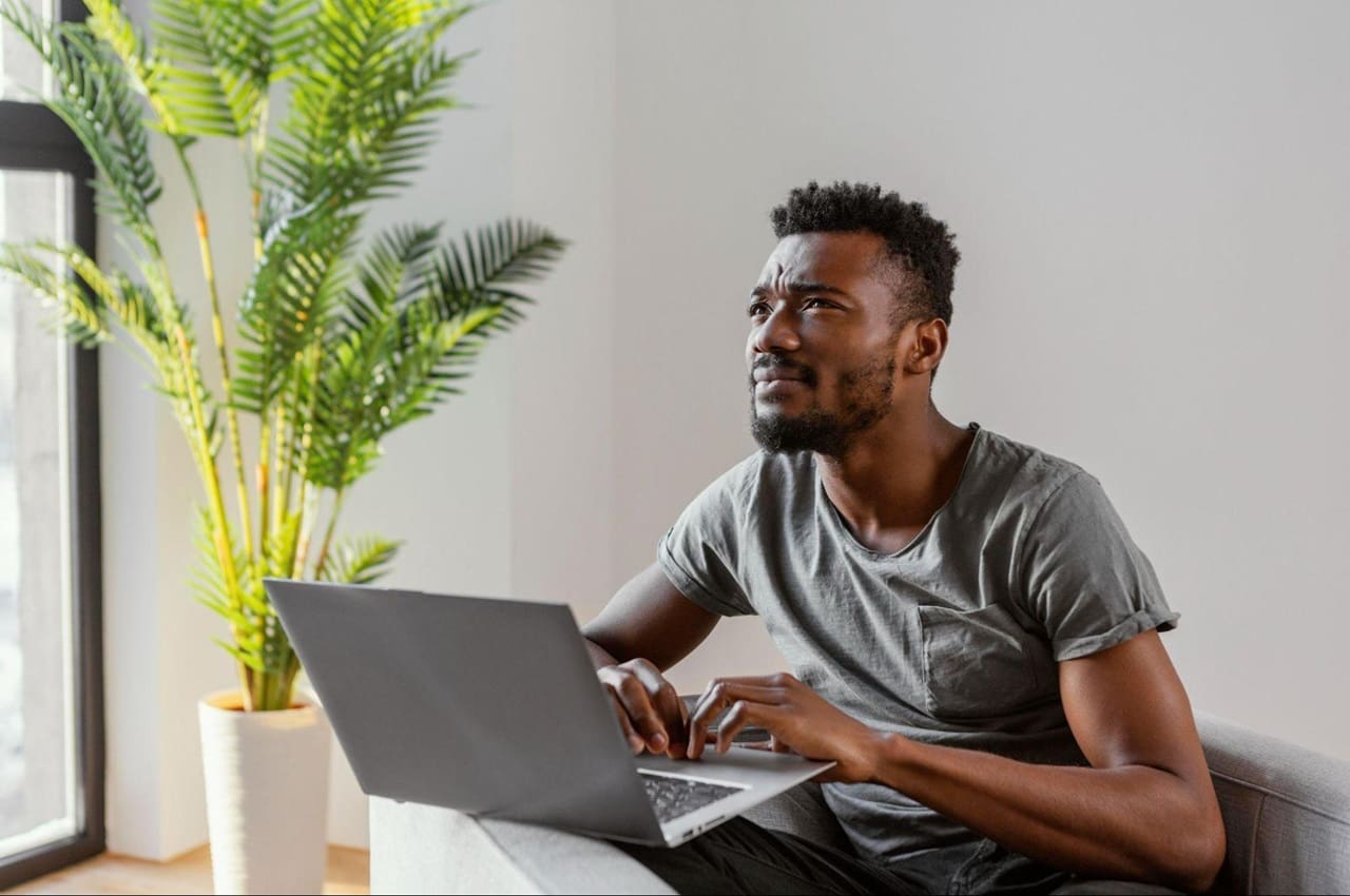 Homem com expressão concentrada digita no laptop do escritório e observa energia luminosa que vem da janela.